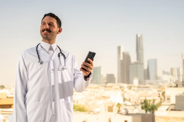 التواصل والدردشة عبر الهاتف المحمول، مفهوم الفحص الطبي، العمل في القطاع الصحي، تقديم الخدمات العلاجية للمرضى, طبيب عربي خليجي سعودي يرتدي الثوب التقليدي والمعطف الطبي يضع السماعة الطبية يعمل في المراكز الصحية والمستشفيات، خلفية أبراج الرياض