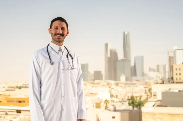 مفهوم الفحص الطبي، العمل في القطاع الصحي، تقديم الخدمات العلاجية للمرضى, طبيب عربي خليجي سعودي يرتدي الثوب التقليدي والمعطف الطبي يضع السماعة الطبية يعمل في المراكز الصحية والمستشفيات، خلفية أبراج الرياض