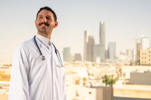 مفهوم الفحص الطبي، العمل في القطاع الصحي، تقديم الخدمات العلاجية للمرضى, طبيب عربي خليجي سعودي يرتدي الثوب التقليدي والمعطف الطبي يضع السماعة الطبية يعمل في المراكز الصحية والمستشفيات، خلفية أبراج الرياض