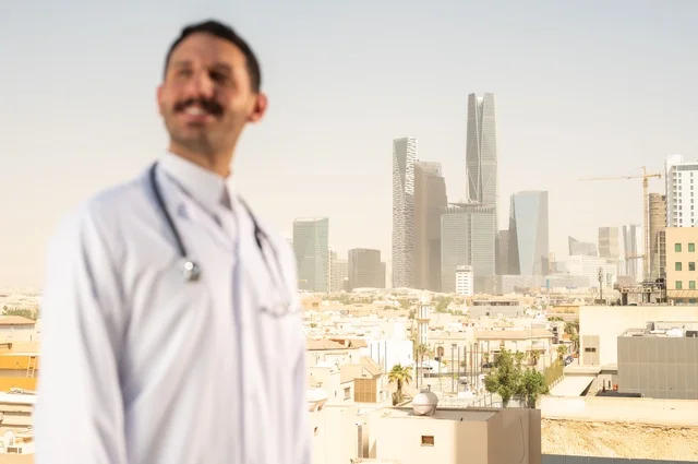 مفهوم الفحص الطبي، العمل في القطاع الصحي، تقديم الخدمات العلاجية للمرضى, طبيب عربي خليجي سعودي يرتدي الثوب التقليدي والمعطف الطبي يضع السماعة الطبية يعمل في المراكز الصحية والمستشفيات، خلفية أبراج الرياض