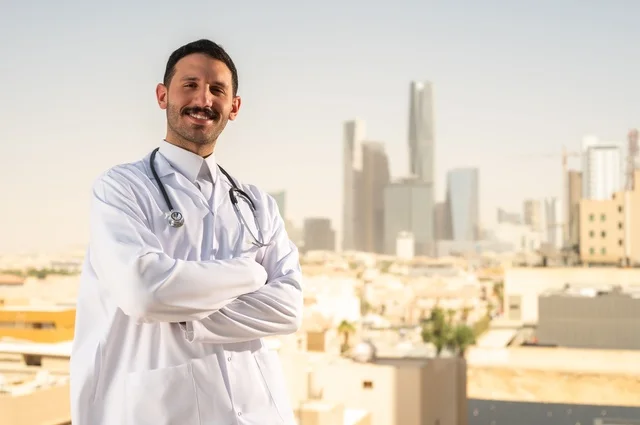 العمل في القطاع الصحي، مفهوم الفحص الطبي، تقديم الخدمات العلاجية للمرضى, طبيب عربي خليجي سعودي يرتدي الثوب التقليدي والمعطف الطبي يضع السماعة الطبية يقف بأيدي متقاطعة يعمل في المراكز الصحية والمستشفيات، خلفية أبراج الرياض
