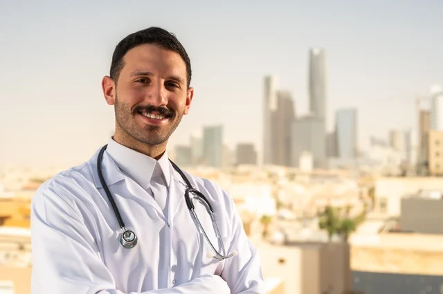 العمل في القطاع الصحي، مفهوم الفحص الطبي، تقديم الخدمات العلاجية للمرضى, طبيب عربي خليجي سعودي يرتدي الثوب التقليدي والمعطف الطبي يضع السماعة الطبية يقف بأيدي متقاطعة يعمل في المراكز الصحية والمستشفيات، خلفية أبراج الرياض