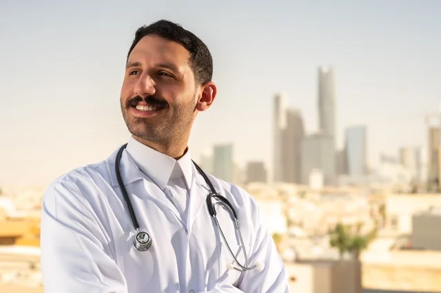 العمل في القطاع الصحي، مفهوم الفحص الطبي، تقديم الخدمات العلاجية للمرضى, طبيب عربي خليجي سعودي يرتدي الثوب التقليدي والمعطف الطبي يضع السماعة الطبية يقف بأيدي متقاطعة يعمل في المراكز الصحية والمستشفيات، خلفية أبراج الرياض