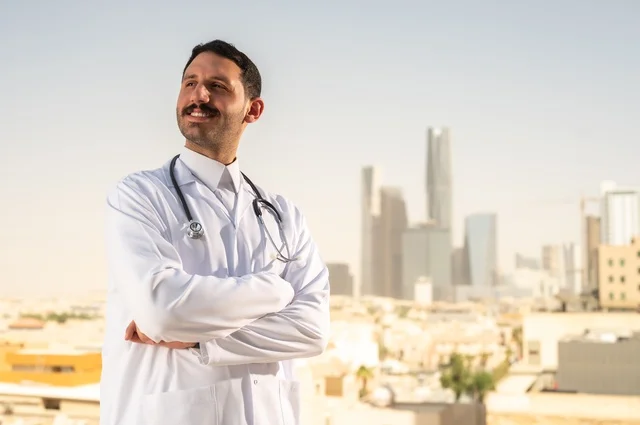 العمل في القطاع الصحي، مفهوم الفحص الطبي، تقديم الخدمات العلاجية للمرضى, طبيب عربي خليجي سعودي يرتدي الثوب التقليدي والمعطف الطبي يضع السماعة الطبية يقف بأيدي متقاطعة يعمل في المراكز الصحية والمستشفيات، خلفية أبراج الرياض