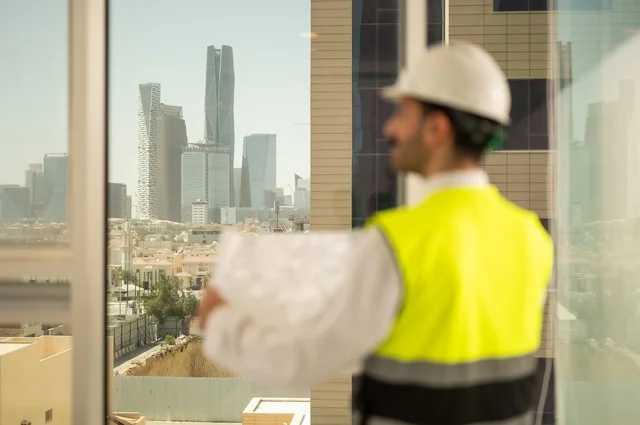 Saudi national construction projects, architectural concept, following up and reviewing the engineering project plan, interest in urban development by Saudi engineers, a Saudi Gulf Arab engineer wearing a work jacket and a white protective helmet performing daily tasks in the office, Riyadh towers background
