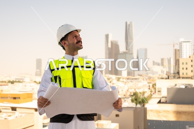 الاهتمام بالتطور العمراني بسواعد المهندسين السعوديين, مهندس عربي خليجي سعودي يرتدي سترة العمل وخوذة الحماية البيضاء ينجز المهام اليومية في المكتب، متابعة مخطط المشروع الهندسي وتدقيقه, مفهوم الهندسة المعمارية، مشاريع إعمارية سعودية وطنية، خلفية أبراج الرياض
