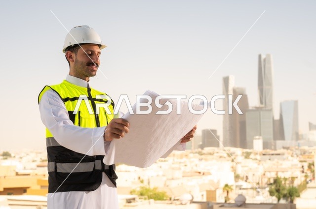 الاهتمام بالتطور العمراني بسواعد المهندسين السعوديين, مهندس عربي خليجي سعودي يرتدي سترة العمل وخوذة الحماية البيضاء ينجز المهام اليومية في المكتب، متابعة مخطط المشروع الهندسي وتدقيقه, مفهوم الهندسة المعمارية، مشاريع إعمارية سعودية وطنية، خلفية أبراج الرياض