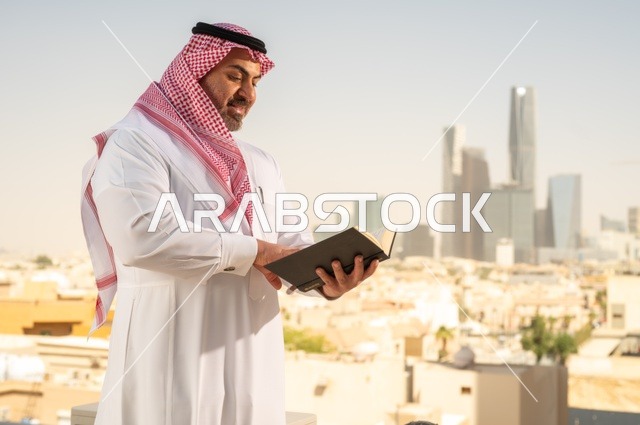 الاستمتاع بممارسة هواية المطالعة، رجل كبير السن عربي خليجي سعودي مبتسم يرتدي الثوب التقليدي والشماغ يقوم بقراءة كتاب بإيماءات السعادة والسرور، مفهوم التثقيف والمعرفة، القصص والروايات الممتعة، خلفية أبراج الرياض