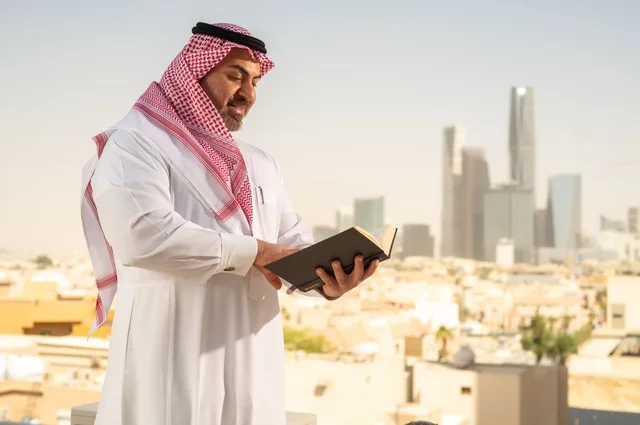 Enjoying reading, an elderly Saudi Arabian Gulf man smiling in traditional thobe and shemagh reading a book with gestures of happiness and pleasure, the concept of education and knowledge, interesting stories and novels, Riyadh towers background