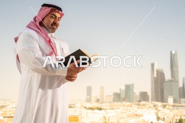 الاستمتاع بممارسة هواية المطالعة، رجل كبير السن عربي خليجي سعودي مبتسم يرتدي الثوب التقليدي والشماغ يقوم بقراءة كتاب بإيماءات السعادة والسرور، مفهوم التثقيف والمعرفة، القصص والروايات الممتعة، خلفية أبراج الرياض