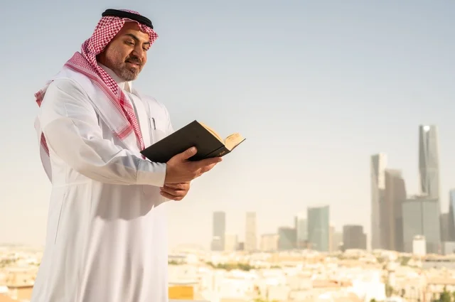 Enjoying reading, an elderly Saudi Arabian Gulf man smiling in traditional thobe and shemagh reading a book with gestures of happiness and pleasure, the concept of education and knowledge, interesting stories and novels, Riyadh towers background