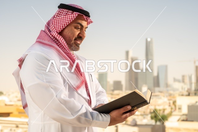 الاستمتاع بممارسة هواية المطالعة، رجل كبير السن عربي خليجي سعودي مبتسم يرتدي الثوب التقليدي والشماغ يقوم بقراءة كتاب بإيماءات السعادة والسرور، مفهوم التثقيف والمعرفة، القصص والروايات الممتعة، خلفية أبراج الرياض