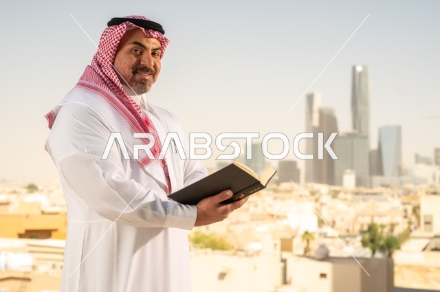 الاستمتاع بممارسة هواية المطالعة، رجل كبير السن عربي خليجي سعودي مبتسم يرتدي الثوب التقليدي والشماغ يقوم بقراءة كتاب بإيماءات السعادة والسرور، مفهوم التثقيف والمعرفة، القصص والروايات الممتعة، خلفية أبراج الرياض