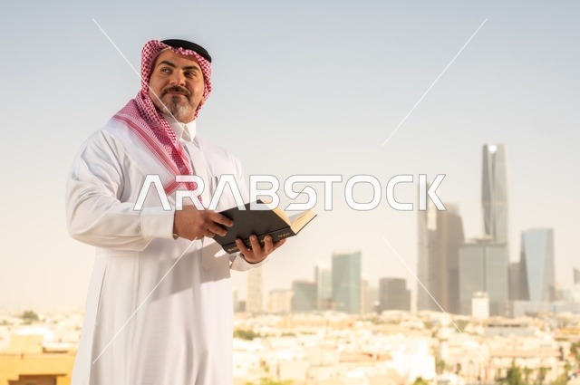 الاستمتاع بممارسة هواية المطالعة، رجل كبير السن عربي خليجي سعودي مبتسم يرتدي الثوب التقليدي والشماغ يقوم بقراءة كتاب بإيماءات السعادة والسرور، مفهوم التثقيف والمعرفة، القصص والروايات الممتعة، خلفية أبراج الرياض