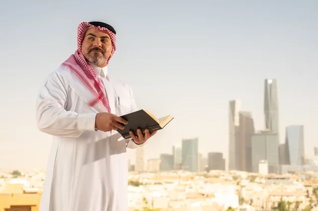 Enjoying reading, an elderly Saudi Arabian Gulf man smiling in traditional thobe and shemagh reading a book with gestures of happiness and pleasure, the concept of education and knowledge, interesting stories and novels, Riyadh towers background