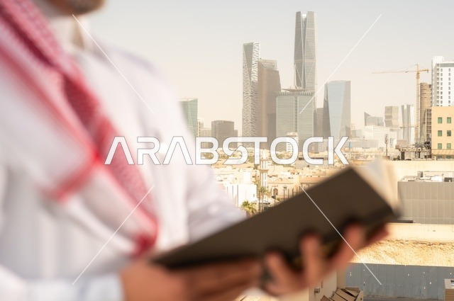 الاستمتاع بممارسة هواية المطالعة، رجل كبير السن عربي خليجي سعودي مبتسم يرتدي الثوب التقليدي والشماغ يقوم بقراءة كتاب بإيماءات السعادة والسرور، مفهوم التثقيف والمعرفة، القصص والروايات الممتعة، خلفية أبراج الرياض