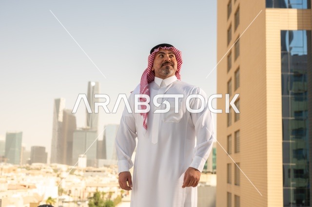 الفخر والاعتزاز بالدولة السعودية، أبراج وناطحات سحاب مدينة الرياض، رجل عربي خليجي سعودي يرتدي الثوب والشماغ التقليدي يقف باستقامة بايماءات السرور، التقدم والتطور المعماري في دول الخليج