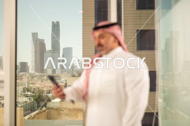 تصفح مواقع التواصل الاجتماعي، جهاز تقني حديث ومتطور،  رجل عربي خليجي سعودي يرتدي الثوب والشماغ التقليدي يقوم باستخدام الهاتف المحمول، إنجاز العمل عن بعد، خلفية الأبراج وناطحات السحاب في مدينة الرياض