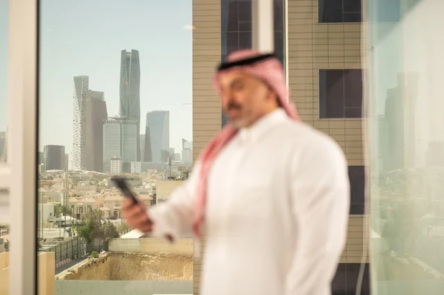تصفح مواقع التواصل الاجتماعي، جهاز تقني حديث ومتطور،  رجل عربي خليجي سعودي يرتدي الثوب والشماغ التقليدي يقوم باستخدام الهاتف المحمول، إنجاز العمل عن بعد، خلفية الأبراج وناطحات السحاب في مدينة الرياض