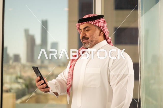تصفح مواقع التواصل الاجتماعي، جهاز تقني حديث ومتطور،  رجل عربي خليجي سعودي يرتدي الثوب والشماغ التقليدي يقوم باستخدام الهاتف المحمول، إنجاز العمل عن بعد، خلفية الأبراج وناطحات السحاب في مدينة الرياض