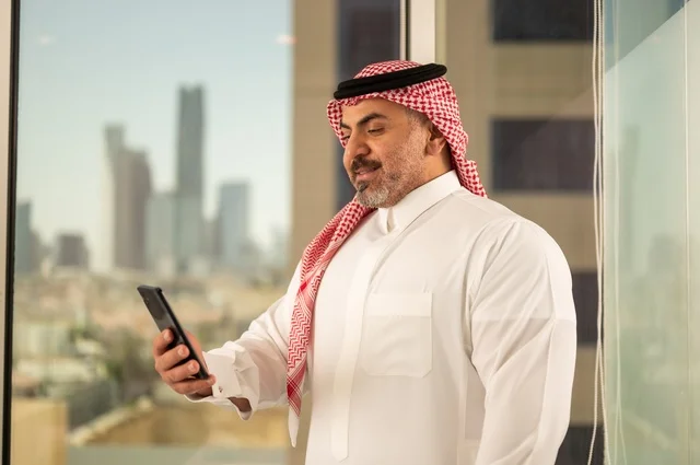 تصفح مواقع التواصل الاجتماعي، جهاز تقني حديث ومتطور،  رجل عربي خليجي سعودي يرتدي الثوب والشماغ التقليدي يقوم باستخدام الهاتف المحمول، إنجاز العمل عن بعد، خلفية الأبراج وناطحات السحاب في مدينة الرياض