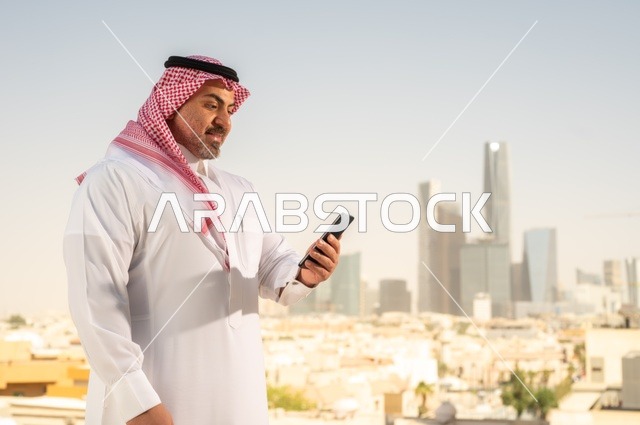 تصفح مواقع التواصل الاجتماعي، جهاز تقني حديث ومتطور،  رجل عربي خليجي سعودي يرتدي الثوب والشماغ التقليدي يقوم باستخدام الهاتف المحمول، إنجاز العمل عن بعد، خلفية الأبراج وناطحات السحاب في مدينة الرياض