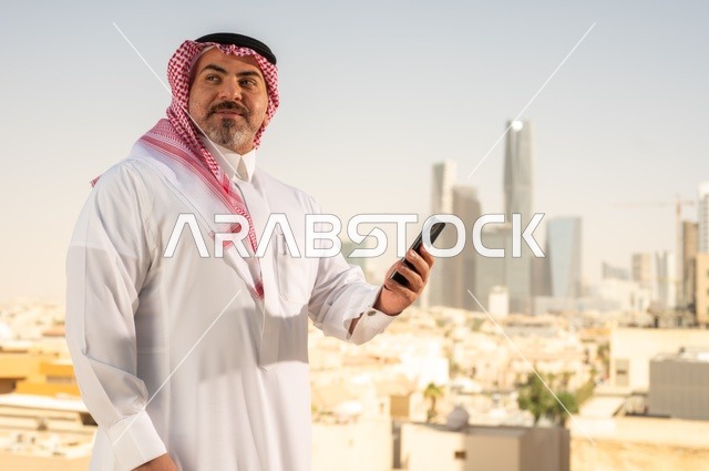 تصفح مواقع التواصل الاجتماعي، جهاز تقني حديث ومتطور،  رجل عربي خليجي سعودي يرتدي الثوب والشماغ التقليدي يقوم باستخدام الهاتف المحمول، إنجاز العمل عن بعد، خلفية الأبراج وناطحات السحاب في مدينة الرياض