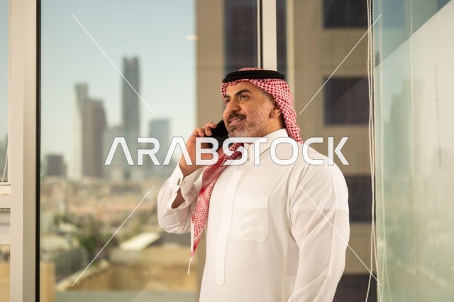 استخدام جهاز تقني حديث ومتطور،  رجل عربي خليجي سعودي يرتدي الثوب التقليدي يقوم بإجراء مكالمة هاتفية عبر الهاتف المحمول، التقدم والتطور المعماري في دول الخليج، خلفية الأبراج وناطحات السحاب في مدينة الرياض