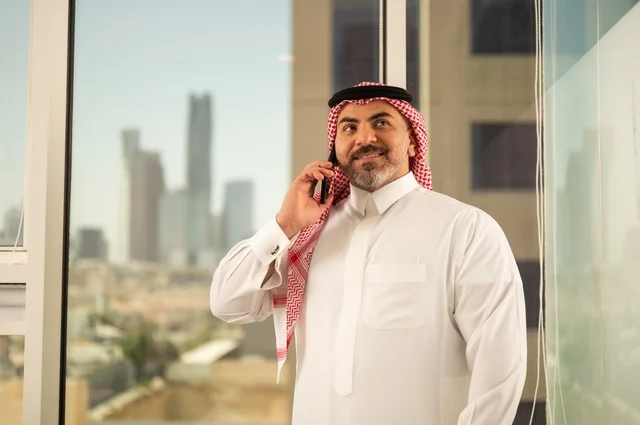 Using a modern and advanced technical device, a close-up shot of a Saudi Arabian Gulf man wearing traditional thobe making a phone call on a mobile phone, the progress and architectural development in the Gulf countries, the background of towers and skyscrapers in the city of Riyadh