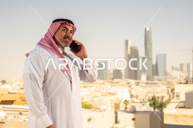 استخدام جهاز تقني حديث ومتطور،  رجل عربي خليجي سعودي يرتدي الثوب التقليدي يقوم بإجراء مكالمة هاتفية عبر الهاتف المحمول، التقدم والتطور المعماري في دول الخليج، خلفية الأبراج وناطحات السحاب في مدينة الرياض