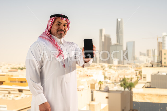 عرض شاشة سوداء فارغة، جهاز تقني حديث ومتطور، رجل عربي خليجي سعودي يرتدي الثوب والشماغ التقليدي يقوم باستخدام الهاتف المحمول، إنجاز العمل عن بعد، خلفية الأبراج وناطحات السحاب في مدينة الرياض