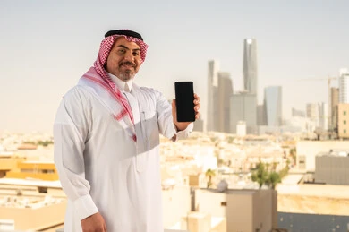 Blank black screen, modern and advanced technical device, Saudi Arabian Gulf man wearing traditional thobe and shemagh using mobile phone, doing remote work, background of towers and skyscrapers in Riyadh