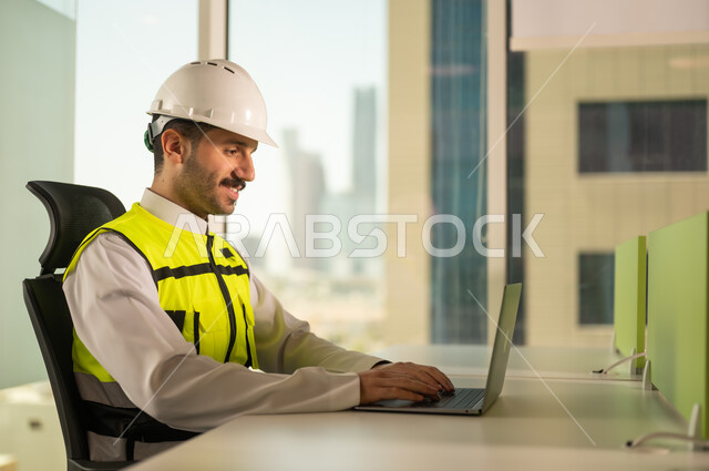 متابعة مخطط المشروع الهندسي اونلاين, مفهوم الهندسة المعمارية، مشاريع إعمارية سعودية وطنية، الاهتمام بالتطور العمراني بسواعد المهندسين السعوديين, مهندس عربي خليجي سعودي يرتدي سترة العمل وخوذة الحماية البيضاء ينجز المهام اليومية عبر الحاسب المحمول