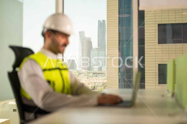 متابعة مخطط المشروع الهندسي اونلاين, مفهوم الهندسة المعمارية، مشاريع إعمارية سعودية وطنية، الاهتمام بالتطور العمراني بسواعد المهندسين السعوديين, مهندس عربي خليجي سعودي يرتدي سترة العمل وخوذة الحماية البيضاء ينجز المهام اليومية عبر الحاسب المحمول
