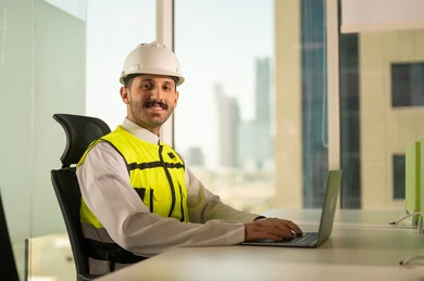 Follow up on the engineering project plan online, the concept of architectural engineering, Saudi national construction projects, interest in urban development through the hands of Saudi engineers, a Saudi Gulf Arab engineer wearing a work jacket and a white protective helmet completing daily tasks via laptop