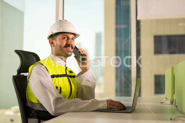 اجراء مكالمة هاتفية عبر الهاتف المحمول, متابعة مخطط المشروع الهندسي اونلاين,  مشاريع إعمارية سعودية وطنية، الاهتمام بالتطور العمراني بسواعد المهندسين السعوديين, مهندس عربي خليجي سعودي يرتدي سترة العمل وخوذة الحماية البيضاء ينجز المهام اليومية عبر الحاسب المحمول