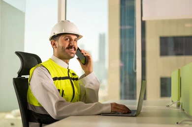Making a phone call via mobile phone, following up on the engineering project plan online, Saudi national construction projects, taking care of urban development with the help of Saudi engineers, a Saudi Gulf Arab engineer wearing a work jacket and a white protective helmet completing daily tasks via laptop
