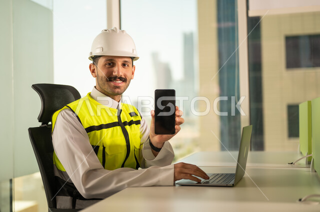 عرض شاشة هاتف محمول سوداء فارغة, متابعة مخطط المشروع الهندسي اونلاين,  مشاريع إعمارية سعودية وطنية، الاهتمام بالتطور العمراني بسواعد المهندسين السعوديين, مهندس عربي خليجي سعودي يرتدي سترة العمل وخوذة الحماية البيضاء ينجز المهام اليومية عبر الحاسب المحمول