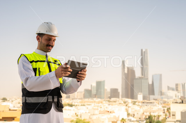 دمج التقنية في الهندسة المعمارية, متابعة مخطط المشروع الهندسي اونلاين, مشاريع إعمارية سعودية وطنية، الاهتمام بالتطور العمراني بسواعد المهندسين السعوديين, مهندس عربي خليجي سعودي يرتدي سترة العمل وخوذة الحماية البيضاء ينجز المهام اليومية عبر جهاز التابلت