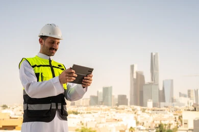 دمج التقنية في الهندسة المعمارية, متابعة مخطط المشروع الهندسي اونلاين, مشاريع إعمارية سعودية وطنية، الاهتمام بالتطور العمراني بسواعد المهندسين السعوديين, مهندس عربي خليجي سعودي يرتدي سترة العمل وخوذة الحماية البيضاء ينجز المهام اليومية عبر جهاز التابلت