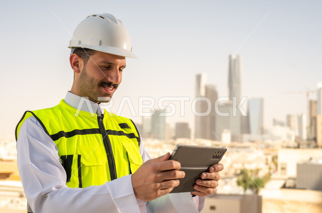 دمج التقنية في الهندسة المعمارية, متابعة مخطط المشروع الهندسي اونلاين, مشاريع إعمارية سعودية وطنية، الاهتمام بالتطور العمراني بسواعد المهندسين السعوديين, مهندس عربي خليجي سعودي يرتدي سترة العمل وخوذة الحماية البيضاء ينجز المهام اليومية عبر جهاز التابلت