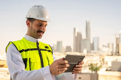 دمج التقنية في الهندسة المعمارية, متابعة مخطط المشروع الهندسي اونلاين, مشاريع إعمارية سعودية وطنية، الاهتمام بالتطور العمراني بسواعد المهندسين السعوديين, مهندس عربي خليجي سعودي يرتدي سترة العمل وخوذة الحماية البيضاء ينجز المهام اليومية عبر جهاز التابلت