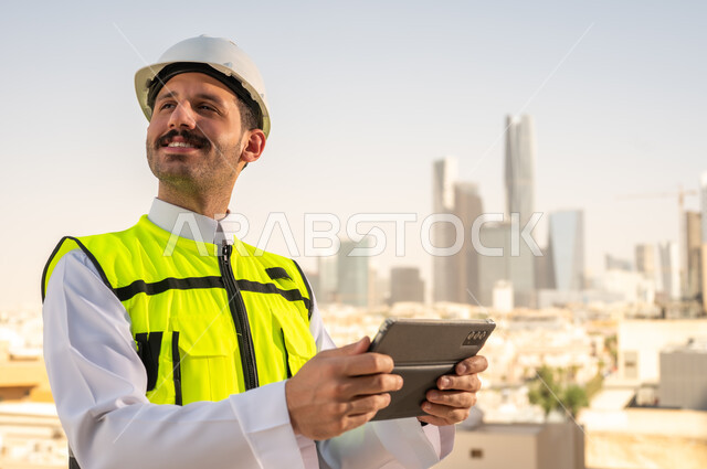 دمج التقنية في الهندسة المعمارية, متابعة مخطط المشروع الهندسي اونلاين, مشاريع إعمارية سعودية وطنية، الاهتمام بالتطور العمراني بسواعد المهندسين السعوديين, مهندس عربي خليجي سعودي يرتدي سترة العمل وخوذة الحماية البيضاء ينجز المهام اليومية عبر جهاز التابلت