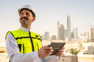 دمج التقنية في الهندسة المعمارية, متابعة مخطط المشروع الهندسي اونلاين, مشاريع إعمارية سعودية وطنية، الاهتمام بالتطور العمراني بسواعد المهندسين السعوديين, مهندس عربي خليجي سعودي يرتدي سترة العمل وخوذة الحماية البيضاء ينجز المهام اليومية عبر جهاز التابلت