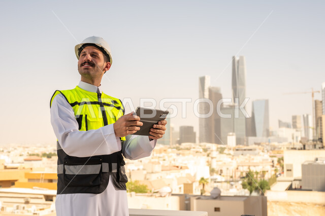 Integrating technology into architecture, following up on engineering project plans online, Saudi national construction projects, focusing on urban development through the efforts of Saudi engineers, a Saudi Gulf Arab engineer wearing a work jacket and a white protective helmet completing daily tasks via a tablet.