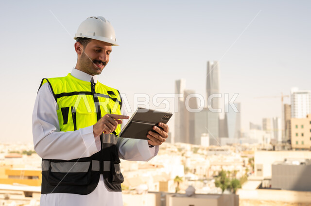 Integrating technology into architecture, following up on engineering project plans online, Saudi national construction projects, focusing on urban development through the efforts of Saudi engineers, a Saudi Gulf Arab engineer wearing a work jacket and a white protective helmet completing daily tasks via a tablet.