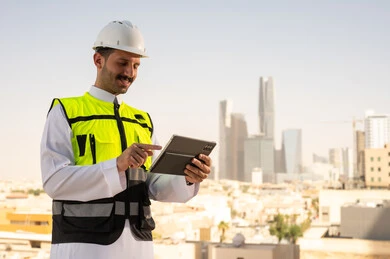 Integrating technology into architecture, following up on engineering project plans online, Saudi national construction projects, focusing on urban development through the efforts of Saudi engineers, a Saudi Gulf Arab engineer wearing a work jacket and a white protective helmet completing daily tasks via a tablet.