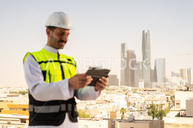دمج التقنية في الهندسة المعمارية, متابعة مخطط المشروع الهندسي اونلاين, مشاريع إعمارية سعودية وطنية، الاهتمام بالتطور العمراني بسواعد المهندسين السعوديين, مهندس عربي خليجي سعودي يرتدي سترة العمل وخوذة الحماية البيضاء ينجز المهام اليومية عبر جهاز التابلت
