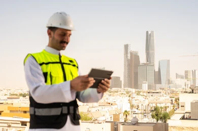 دمج التقنية في الهندسة المعمارية, متابعة مخطط المشروع الهندسي اونلاين, مشاريع إعمارية سعودية وطنية، الاهتمام بالتطور العمراني بسواعد المهندسين السعوديين, مهندس عربي خليجي سعودي يرتدي سترة العمل وخوذة الحماية البيضاء ينجز المهام اليومية عبر جهاز التابلت