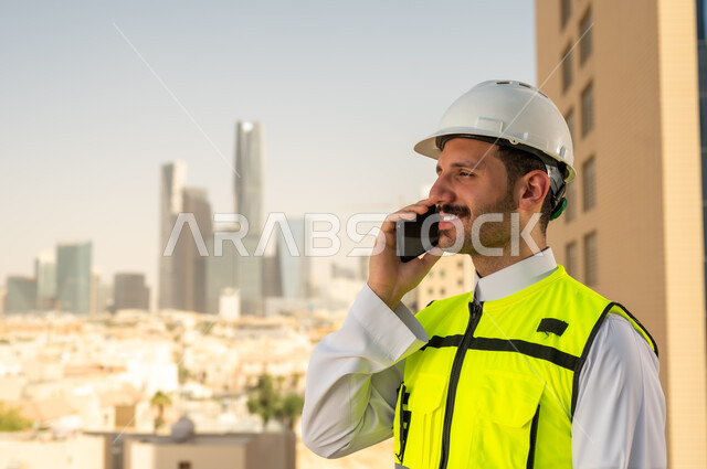 إجراء مكالمة هاتفية, مشاريع إعمارية سعودية وطنية، الاهتمام بالتطور العمراني بسواعد المهندسين السعوديين, مهندس عربي خليجي سعودي يرتدي سترة العمل وخوذة الحماية البيضاء يمسك بيده الهاتف المحمول, دمج التقنية في الهندسة المعمارية, متابعة مخطط المشروع الهندسي